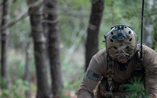 A U.S. military special operations medic in full tactical gear is providing combat casualty care to a downed soldier lying on the ground in a wooded area. The medic is wearing a multicam helmet with face protection, tactical vest, and gloves while attending to what appears to be simulated wounds on the casualty. This appears to be a training exercise based on the controlled environment and use of simulated injury props.