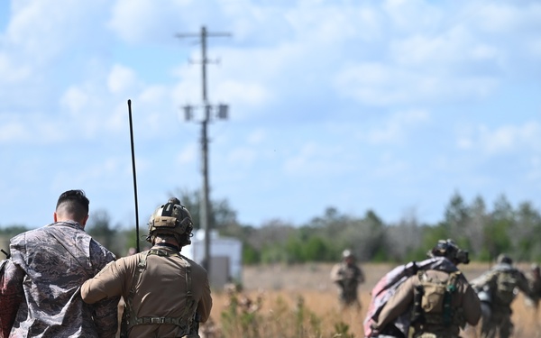 The image shows military personnel in a field environment, with what appears to be a combat search and rescue or personnel recovery exercise. A tactical operator in full gear with a radio antenna is escorting a simulated casualty or role-player in civilian/camouflage clothing across an open grassy field. In the background, additional personnel are conducting similar escort or security activities.