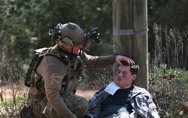 A fully equipped military or special operations soldier is attending to a simulated casualty during what appears to be a training exercise. The seated individual has theatrical makeup applied to simulate injuries and has a triage card attached to their uniform. The responder is performing a head assessment on the mock casualty near a utility pole in a wooded outdoor environment.