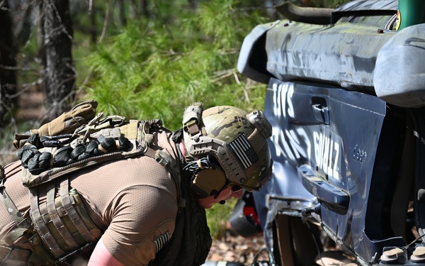 A military or tactical operator in full combat gear is providing medical assistance or extracting a downed personnel near what appears to be a crashed or overturned vehicle. The scene takes place in a wooded outdoor environment, suggesting a training exercise or simulated emergency scenario. The operator is wearing multicam camouflage, a tactical vest, and a helmet with a US flag patch.