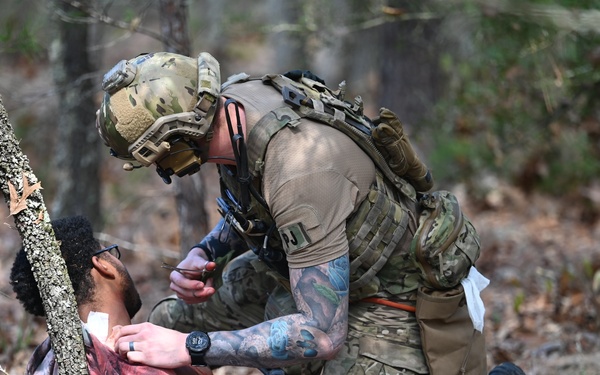 A fully equipped military or tactical operator in multicam camouflage gear is kneeling over a simulated casualty on a forest floor covered in dead leaves. The operator appears to be performing tactical combat casualty care (TCCC) or medical aid on the downed individual, who is wearing civilian-style clothing with visible simulated wound markings. The setting and training indicators suggest this is a military medical training exercise.