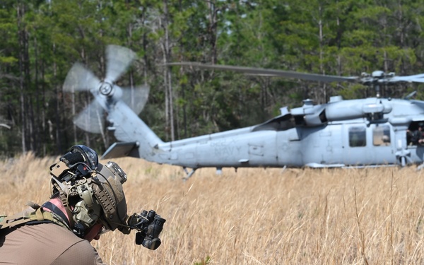 A military training or combat search and rescue (CSAR) exercise is underway in a field clearing surrounded by trees. A heavily equipped special operations soldier wearing a tactical helmet with night vision device mounts is attending to personnel lying on the ground, likely simulated casualties. A gray military helicopter, consistent with an MH-60 Black Hawk variant, hovers or sits on the ground in the background.