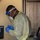 A military medical personnel member wearing full PPE (personal protective equipment) is writing or documenting information at a workstation. The individual is dressed in U.S. Army multicam/OCP camouflage pants beneath a yellow isolation gown, with a face shield, N95 mask, and blue nitrile gloves. The setting appears to be a medical screening or testing station, likely during the COVID-19 pandemic.