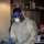 A healthcare worker wearing full personal protective equipment (PPE) is handling what appears to be a specimen vial near a red-and-white cooler/specimen transport container. The individual is dressed in a yellow isolation gown, blue nitrile gloves, N95 respirator mask, safety glasses, and a blue face shield. The setting appears to be an indoor clinical or testing environment with shelving and supplies visible in the background.