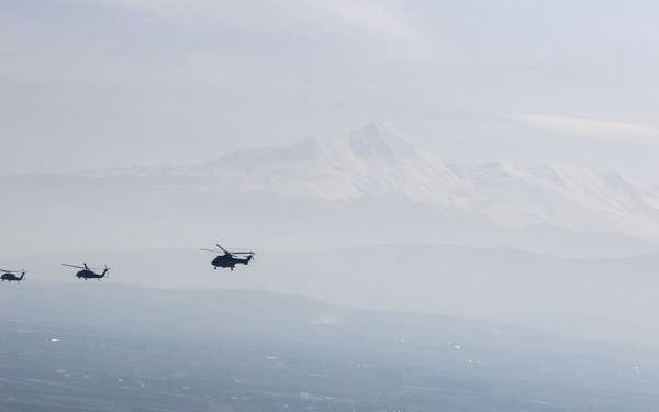 A formation of approximately five military helicopters flying in a loose line formation at altitude over a wide agricultural valley or plain. A large snow-capped mountain is visible in the background through hazy atmospheric conditions. The helicopters appear to be medium/heavy transport types based on their silhouette profiles.
