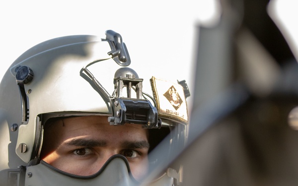 A military aviator or pilot is wearing a flight helmet with an oxygen mask and helmet-mounted equipment attachments. The image is taken from a low angle through foreground aircraft or cockpit structures, creating a dramatic framing effect. The subject appears to be in or near an aircraft, likely during pre-flight or operational activity.