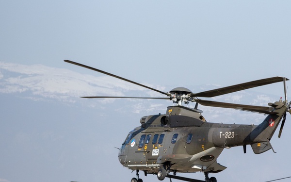 Three military helicopters are flying in formation over a landscape with snow-capped mountains visible in the background. The lead helicopter, marked 'T-323' with a Swiss cross insignia, appears to be an Aerospatiale AS332 Super Puma, while the two lower aircraft appear to be Sikorsky UH-60 Black Hawks. One of the Black Hawks bears a red cross marking, indicating a medical/medevac role.