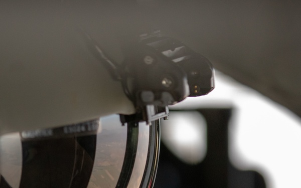 A close-up photograph of a military flight helmet with a tinted visor in the raised/flipped-up position. The visor reflects an aerial landscape view, suggesting the photograph was taken aboard an aircraft in flight. The shallow depth of field keeps the helmet visor in focus while the background remains blurred.
