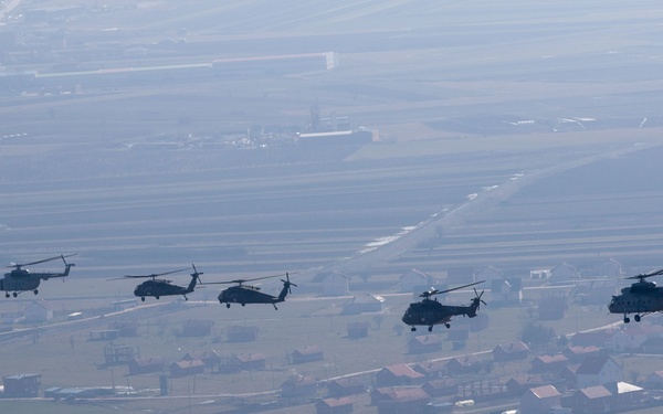 An aerial photograph shows a formation of five military helicopters flying in a loose echelon formation over a rural European landscape. The terrain below features agricultural fields, small villages with red-roofed buildings, and what appears to be an airfield or runway visible in the background. The image was taken from another aircraft, suggesting a coordinated multi-aircraft operation or exercise.