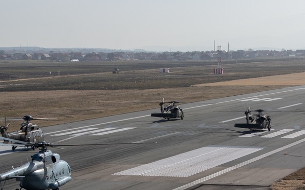 Multiple military helicopters are lined up on an airfield runway, appearing to be preparing for takeoff or conducting taxi operations. The formation includes what appear to be Mi-17 type helicopters in light blue/grey livery in the foreground and darker UH-60 Black Hawk type helicopters in the middle distance. A civilian settlement is visible on the horizon beyond the airfield perimeter.