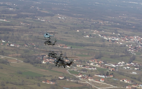 A formation of four military helicopters flying in loose echelon formation over a rural European landscape with scattered villages, agricultural fields, and tree lines. The lead helicopter is a light blue/grey painted large helicopter with the number '301' visible on its fuselage, followed by darker olive/camouflage painted helicopters behind it. The terrain below suggests a Balkan or Eastern European setting based on the architectural style of the buildings.