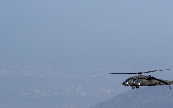 A military helicopter, identifiable as a UH-60 Black Hawk variant with a red cross medical marking, is flying over a mountainous and hazy valley landscape. The aircraft appears to be on a medevac or medical evacuation mission based on the visible red cross insignia on its fuselage. The terrain below shows rolling hills and a broad valley extending into the distance under hazy atmospheric conditions.