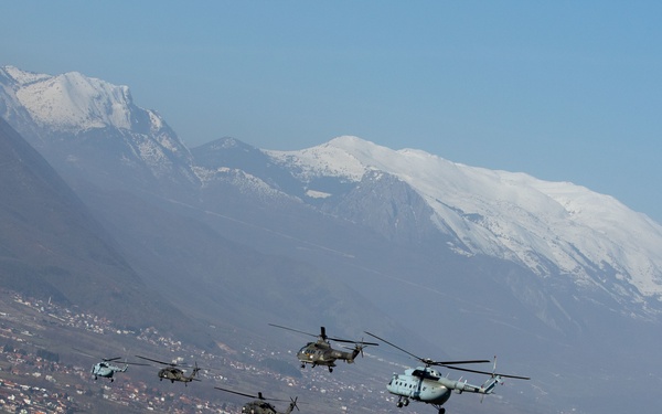A formation of five military helicopters flying over a populated valley town with snow-capped mountains in the background. The helicopters appear to be a mix of types, including what appears to be Soviet/Russian-designed Mi-8/Mi-17 variants and Western-type helicopters, suggesting a multinational or joint military exercise. The scene is set in what appears to be a Balkan or Eastern European mountainous region during winter or early spring.