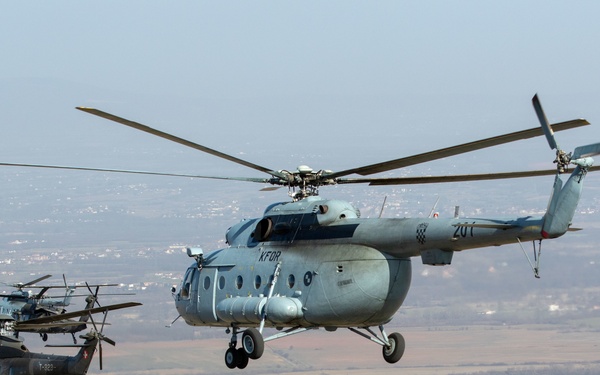 A formation of four military helicopters flying in close proximity over a rural landscape. The lead aircraft is a large Soviet-designed Mi-8/Mi-17 transport helicopter marked with 'KFOR' insignia and tail number '201', accompanied by what appear to be UH-60 Black Hawk helicopters in the background. The scene suggests a multinational NATO KFOR (Kosovo Force) training or patrol mission.