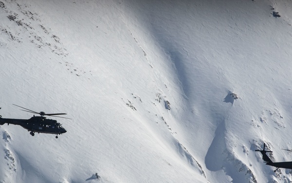 Two military helicopters are flying in formation against a steep, snow-covered mountain face. The helicopters appear dark/black in silhouette against the bright white snow backdrop, creating strong visual contrast. The mountainous terrain shows dramatic ridgelines, shadows, and avalanche chutes across the snow-covered slopes.
