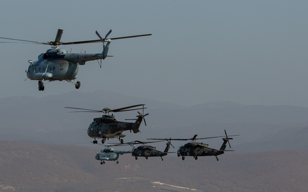 A formation of four military helicopters flying over a mid-sized urban settlement surrounded by hilly terrain. The helicopters appear to represent multiple nations or branches, suggesting a multinational military exercise or patrol mission. The landscape below features a densely built town with residential and commercial structures set against a backdrop of rolling mountains.