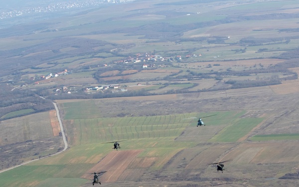 An aerial photograph shows a formation of five military helicopters flying low over a rural, agricultural landscape. The helicopters appear to be of mixed types, flying in a loose echelon or staggered formation. The terrain below consists of patchwork farmland, open fields, and a small settlement visible in the background.