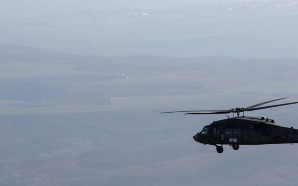 A dark military helicopter marked 'KFOR' is captured in flight over a wide, hazy valley landscape. The aircraft appears to be a UH-60 Black Hawk type helicopter operating under NATO's Kosovo Force (KFOR) mission. The terrain below shows agricultural fields, scattered settlements, and rolling hills under overcast conditions.