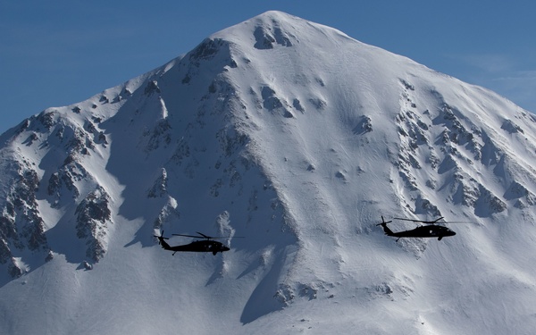 Two military helicopters fly in formation against a dramatic backdrop of a large, heavily snow-covered mountain peak under a clear blue sky. The helicopters appear as dark silhouettes contrasting sharply against the bright white snow-covered slopes. The scene suggests a military training or patrol operation in a high-altitude alpine environment.