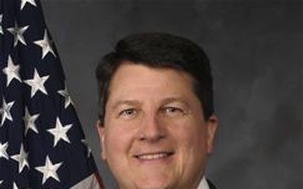 A formal official portrait of a middle-aged man in professional attire, posed in front of an American flag against a neutral gray background. The composition and styling are consistent with a government or political official headshot. The subject is smiling and facing the camera directly.