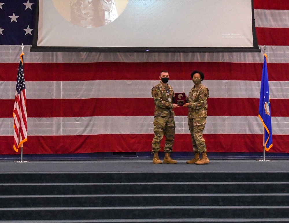 Two military personnel in camouflage uniforms and face masks stand on a stage during what appears to be an awards ceremony. One individual is presenting or receiving a red velvet plaque/award. The stage is decorated with a large American flag backdrop, a US flag on the left, and what appears to be an Air Force flag on the right.