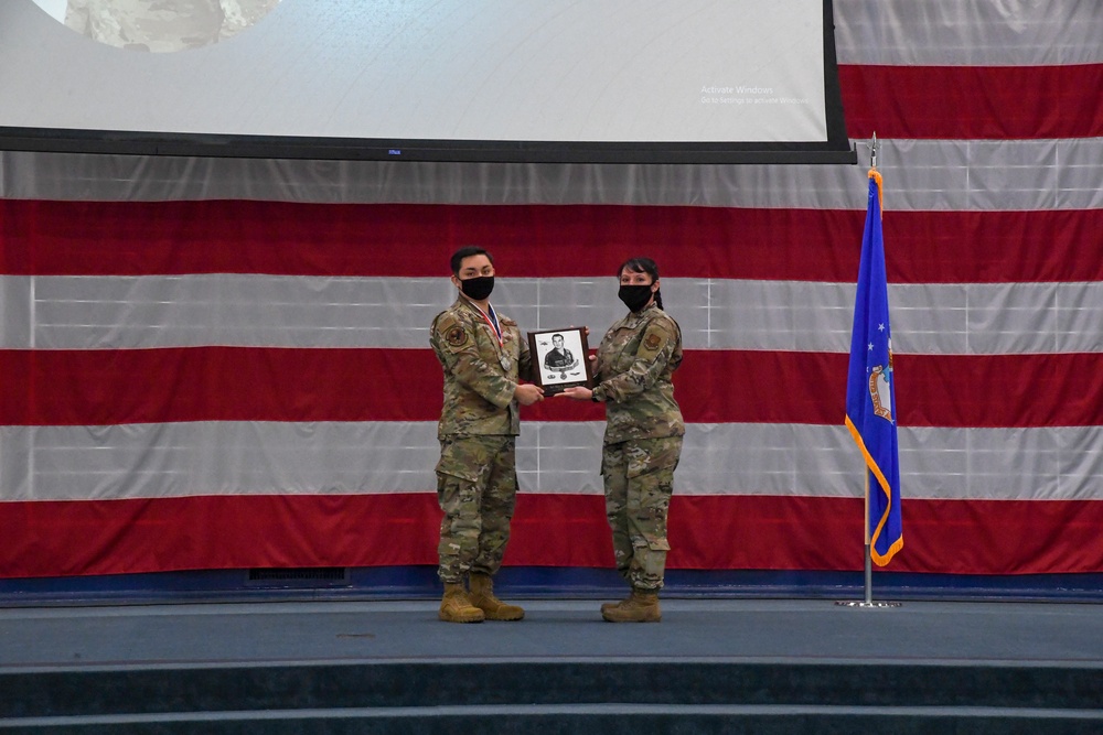 Two military personnel in OCP (Operational Camouflage Pattern) uniforms and black face masks stand on a stage during a formal ceremony. They are jointly holding a framed plaque or award containing a black-and-white portrait photograph. The backdrop features a large American flag and a blue U.S. Air Force flag on a stand.