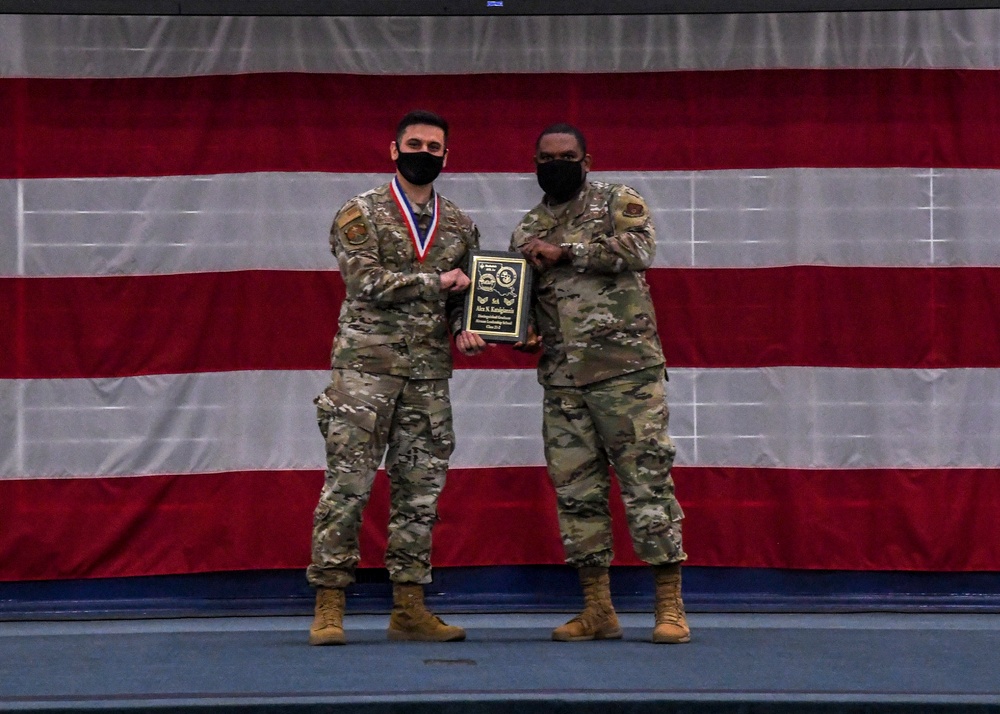 Two military personnel in camouflage uniforms stand together on a stage in front of a large American flag backdrop, jointly holding a plaque during what appears to be an awards ceremony. The individual on the left is wearing a red, white, and blue medal around their neck, indicating they are the award recipient. Both individuals are wearing black face masks, suggesting this event took place during the COVID-19 pandemic period.