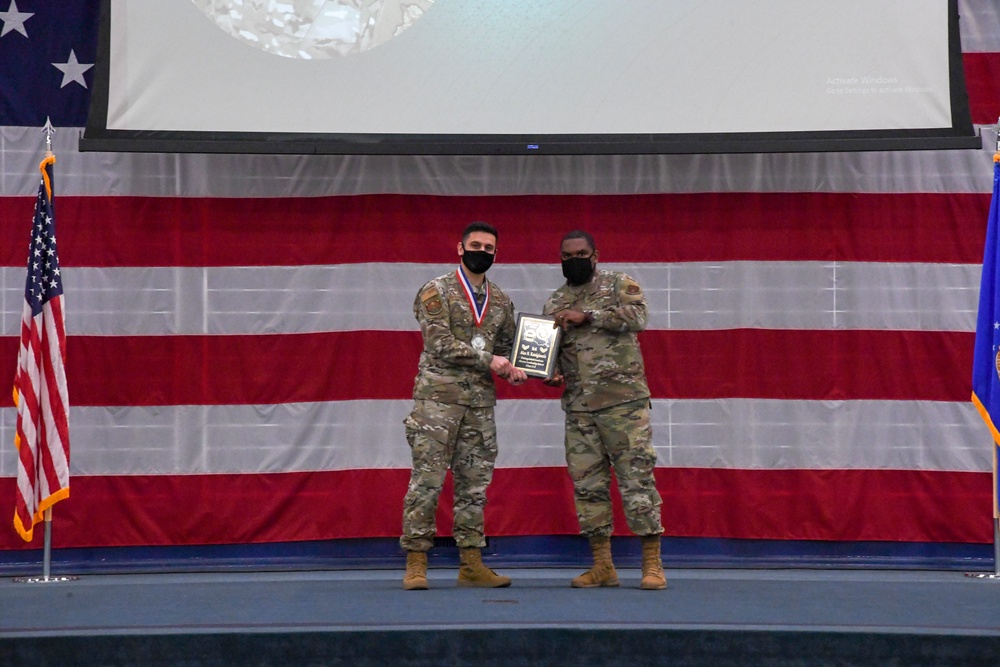 Two military personnel in camouflage uniforms stand on a stage in front of a large American flag backdrop during what appears to be an awards ceremony. The individual on the left is wearing a medal around their neck and both are jointly holding a plaque. Both individuals are wearing black face masks, suggesting this event took place during the COVID-19 pandemic.