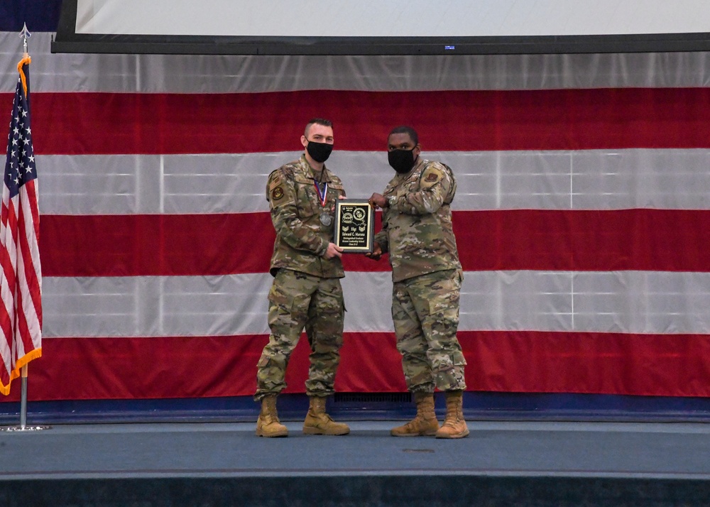 Two military personnel in OCP (Operational Camouflage Pattern) uniforms stand on a stage in front of a large American flag, jointly holding a black award plaque. Both individuals are wearing black face masks, suggesting this ceremony took place during the COVID-19 pandemic period. The scene depicts a formal military awards or recognition ceremony.