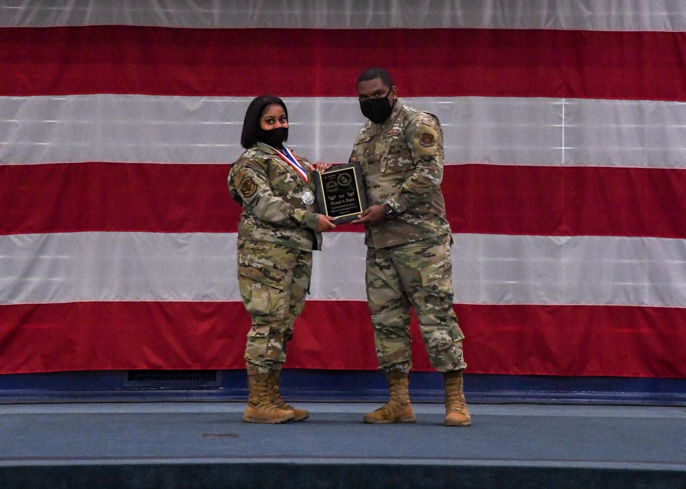 Two military personnel in OCP (Operational Camouflage Pattern) uniforms stand on a stage in front of a large American flag backdrop. They are jointly holding a black commemorative plaque during what appears to be a formal awards ceremony. The service member on the left is also wearing a medal around their neck.