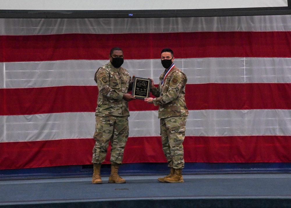 Two military personnel in OCP (Operational Camouflage Pattern) uniforms are standing on a stage in front of a large American flag, jointly holding a black award plaque. The individual on the right is wearing a medal around their neck, suggesting this is an awards or recognition ceremony. Both individuals are wearing black face masks, indicating this event likely took place during the COVID-19 pandemic period.