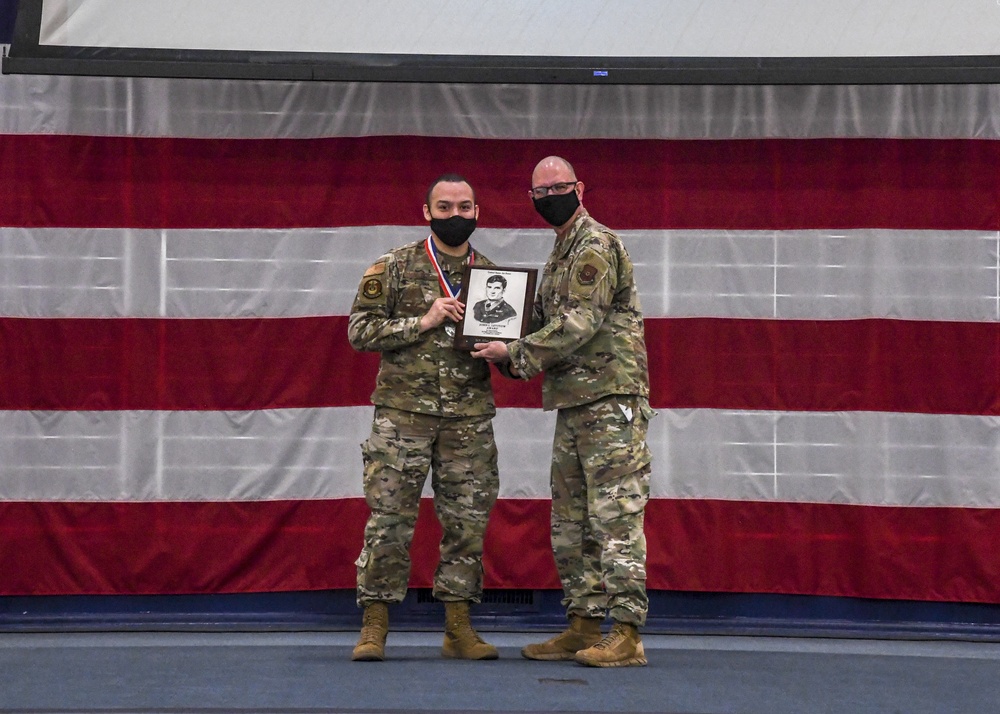 Two military personnel in OCP (Operational Camouflage Pattern) uniforms stand together on a stage in front of a large American flag backdrop, both wearing black face masks. They are jointly holding a framed plaque that appears to contain a portrait photograph and text. The individual on the left is also wearing a medal with a red, white, and blue ribbon, suggesting this is an awards or recognition ceremony.