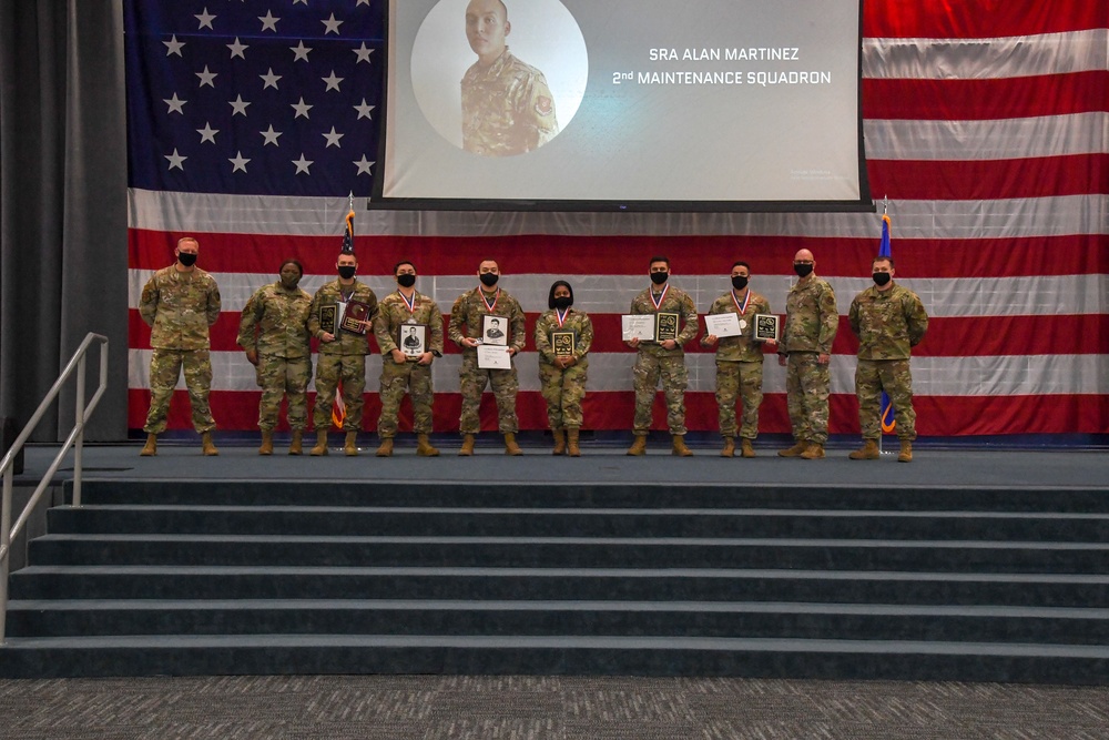A group of approximately ten military personnel in camouflage uniforms pose on a stage during an awards ceremony. Several individuals are holding plaques, certificates, and medals, with a large projection screen displaying 'SRA Alan Martinez, 2nd Maintenance Squadron' behind them. A large American flag serves as a backdrop, and all personnel are wearing face masks.