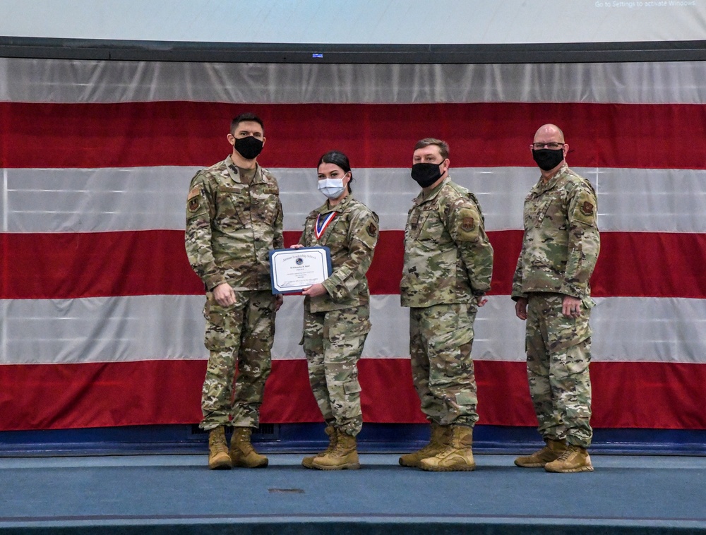 Four military personnel in OCP (Operational Camouflage Pattern) uniforms are posed in front of a large American flag backdrop. One individual, appearing to be female and wearing a medal/ribbon around her neck, is holding a certificate or award document. This appears to be a formal military awards or recognition ceremony.