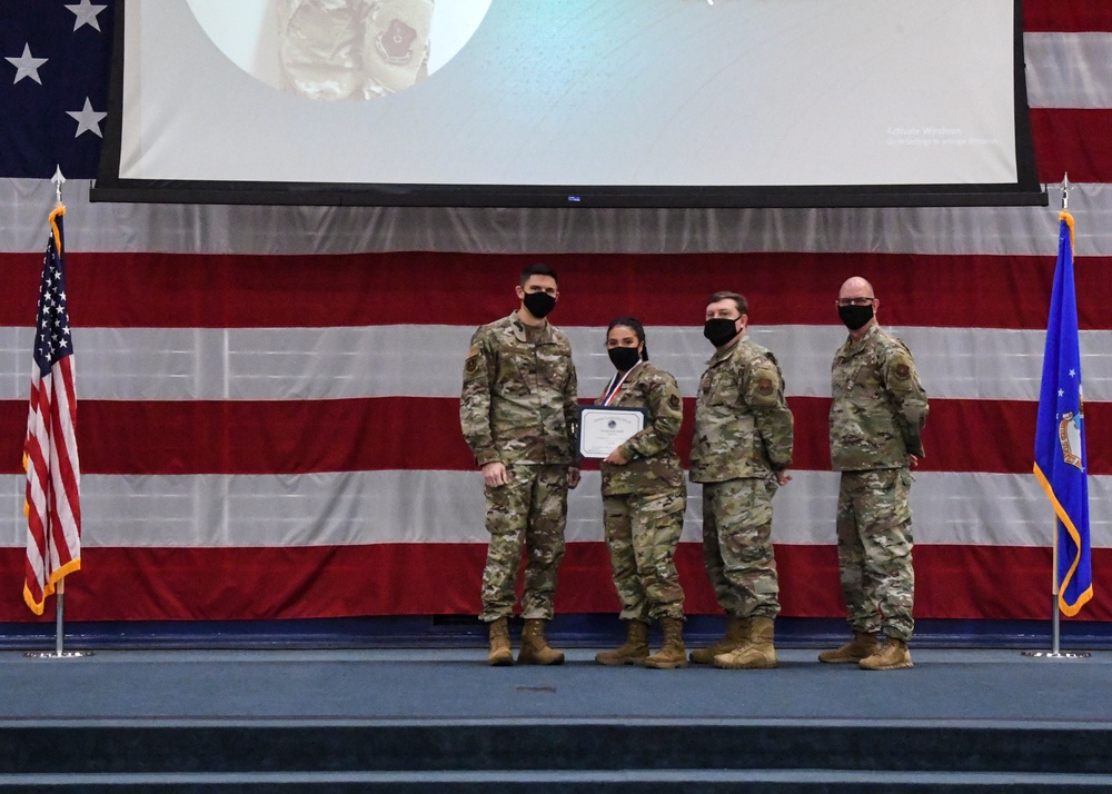 Four military personnel in OCP (Operational Camouflage Pattern) uniforms are posed on a stage during a formal awards ceremony. One individual, appearing to be female, is holding a certificate and wearing a medal around her neck, indicating she is the award recipient. All four individuals are wearing black face masks, suggesting this ceremony took place during the COVID-19 pandemic period.
