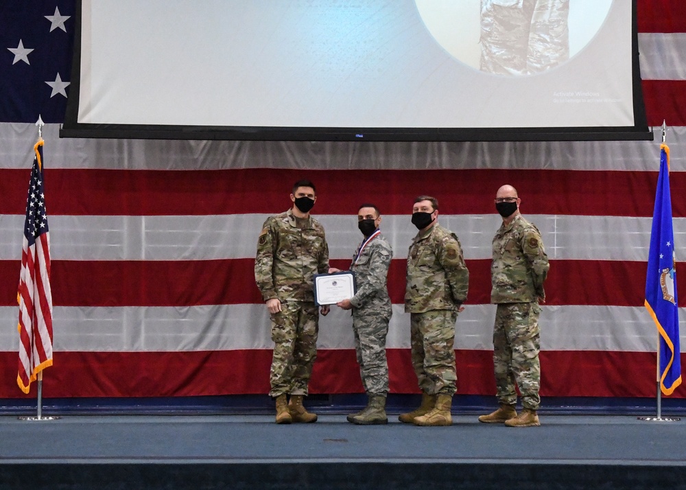 Four military personnel in OCP (Operational Camouflage Pattern) uniforms stand on a stage during a formal awards or recognition ceremony. One individual, wearing a medal and holding a certificate, appears to be the recipient of an award. All four individuals are wearing black face masks, consistent with COVID-19 era protocols.