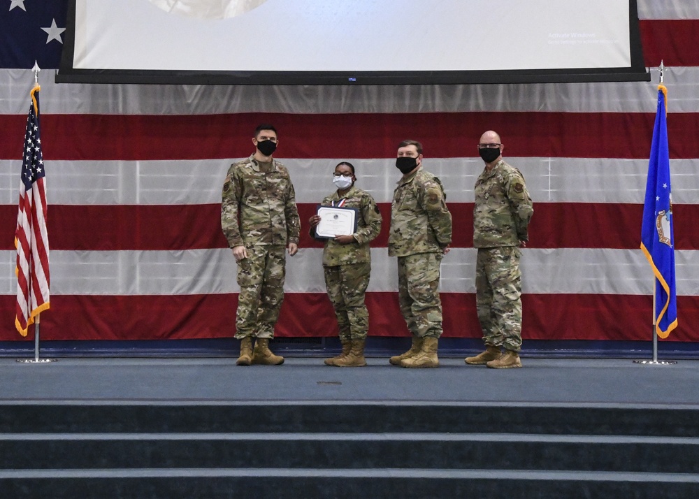Four military personnel in OCP (Operational Camouflage Pattern) uniforms stand on a stage during a formal ceremony. One individual, appearing to be the recipient, holds a certificate/award and is wearing a medal. All four individuals are wearing black face masks, suggesting this ceremony took place during the COVID-19 pandemic period.