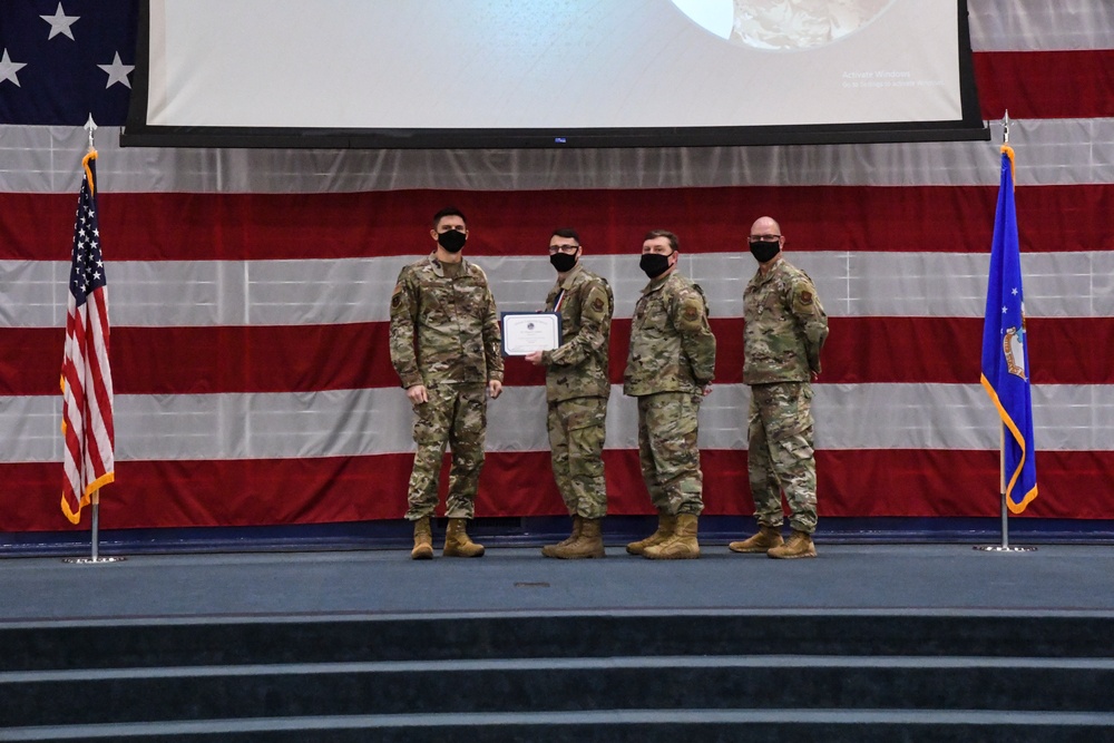 Four military personnel in OCP (Operational Camouflage Pattern) uniforms stand on a stage during a formal ceremony, with two individuals holding a certificate or award document. All four are wearing black face masks, suggesting this event took place during the COVID-19 pandemic period. The backdrop features a large American flag drape and a projection screen, with a US flag on the left and what appears to be a US Air Force flag on the right.