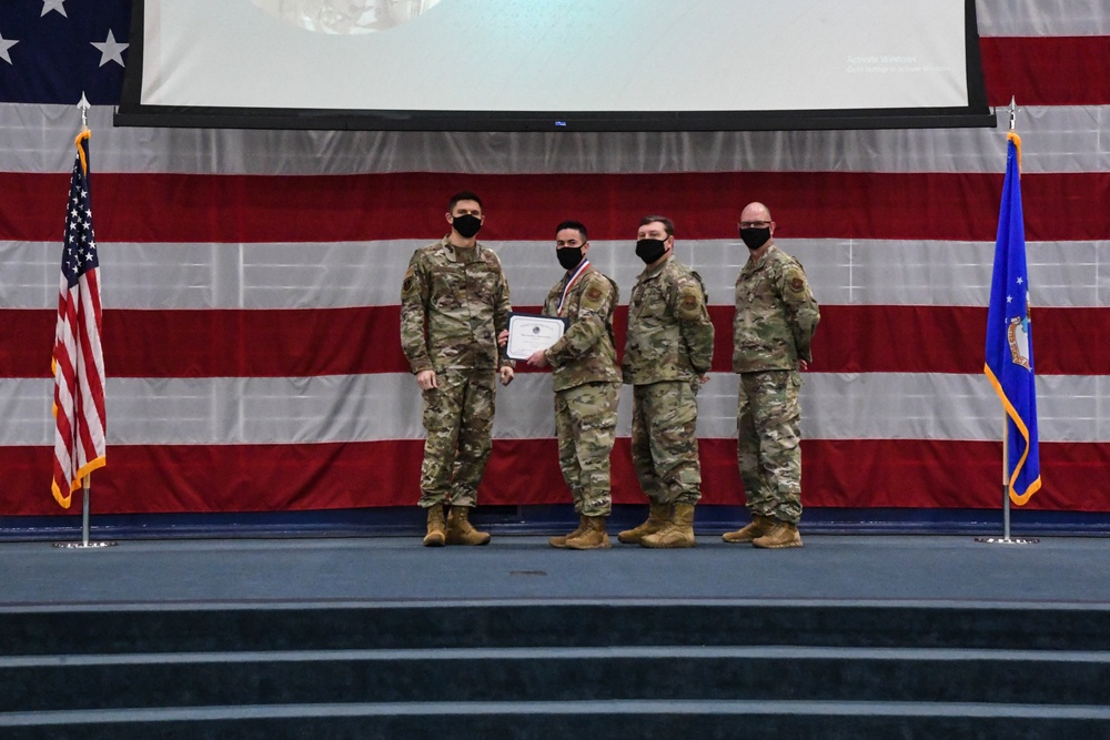 Four military personnel in camouflage uniforms and black face masks stand on a stage during a formal ceremony. One individual wearing a medal is receiving a certificate from another service member, while two others stand at attention. The backdrop features a large American flag and a blue Air Force flag, indicating this is a U.S. Air Force recognition or awards ceremony.