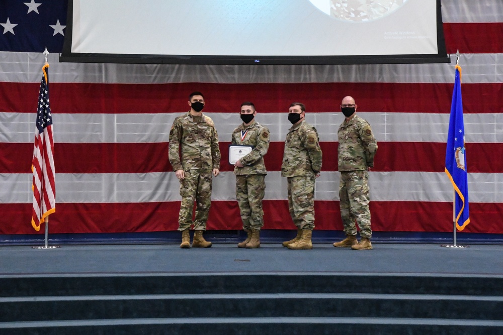 Four military personnel in OCP (Operational Camouflage Pattern) uniforms stand on a stage during what appears to be an awards ceremony. One individual is holding a certificate and wearing a medal around their neck, suggesting they are the recipient of a commendation. All four individuals are wearing black face masks, indicating this event took place during the COVID-19 pandemic period.