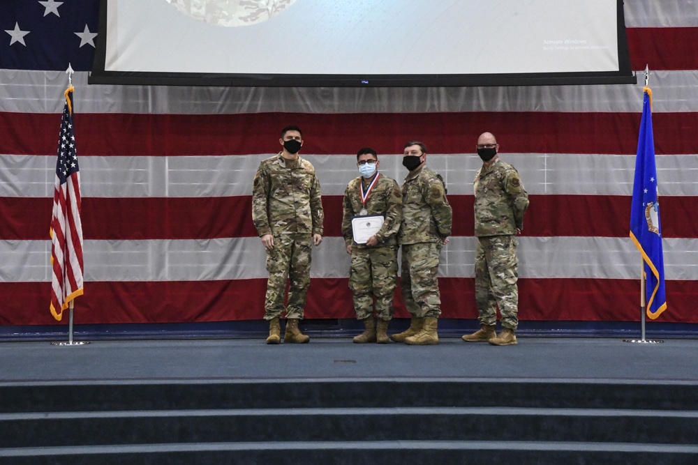 Four military personnel in camouflage uniforms stand on a stage during what appears to be an awards ceremony. The central figure is holding a certificate and wearing a medal around their neck, flanked by three other service members. All four individuals are wearing face masks, suggesting this event took place during the COVID-19 pandemic.