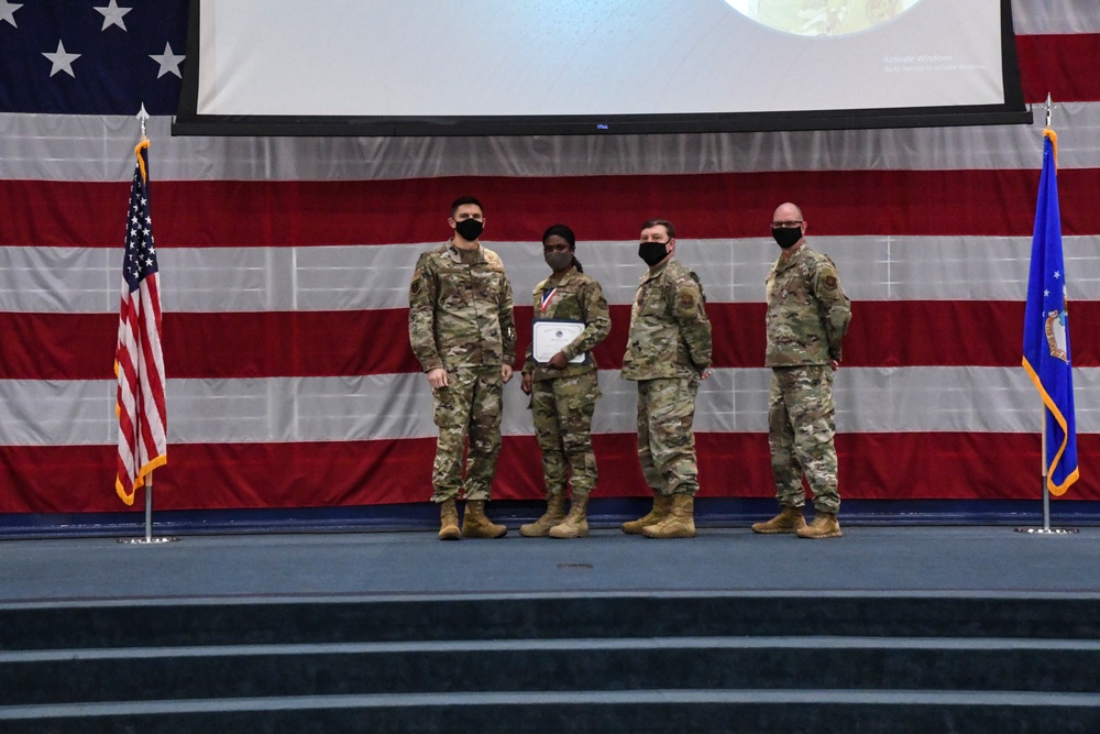 Four military personnel in OCP (Operational Camouflage Pattern) uniforms stand on a stage during what appears to be an awards ceremony. One individual is holding a certificate and wearing a medal around their neck. All four are wearing black face masks, suggesting this event took place during the COVID-19 pandemic.