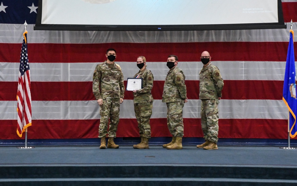 Four military personnel in OCP (Operational Camouflage Pattern) uniforms stand on a stage during a formal ceremony. Two individuals in the center are holding a framed certificate or award document together, suggesting a presentation or recognition event. All four individuals are wearing black face masks, indicating this ceremony took place during the COVID-19 pandemic period.