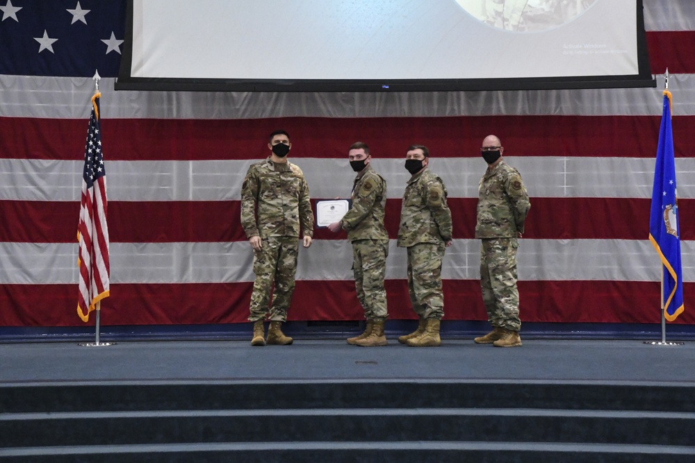 Four military personnel in camouflage uniforms and black face masks are standing on a stage during a formal ceremony. Two individuals in the center are shaking hands while exchanging what appears to be a certificate or award. The backdrop features a large American flag and a blue Air Force flag, suggesting this is a U.S. Air Force recognition or awards ceremony.