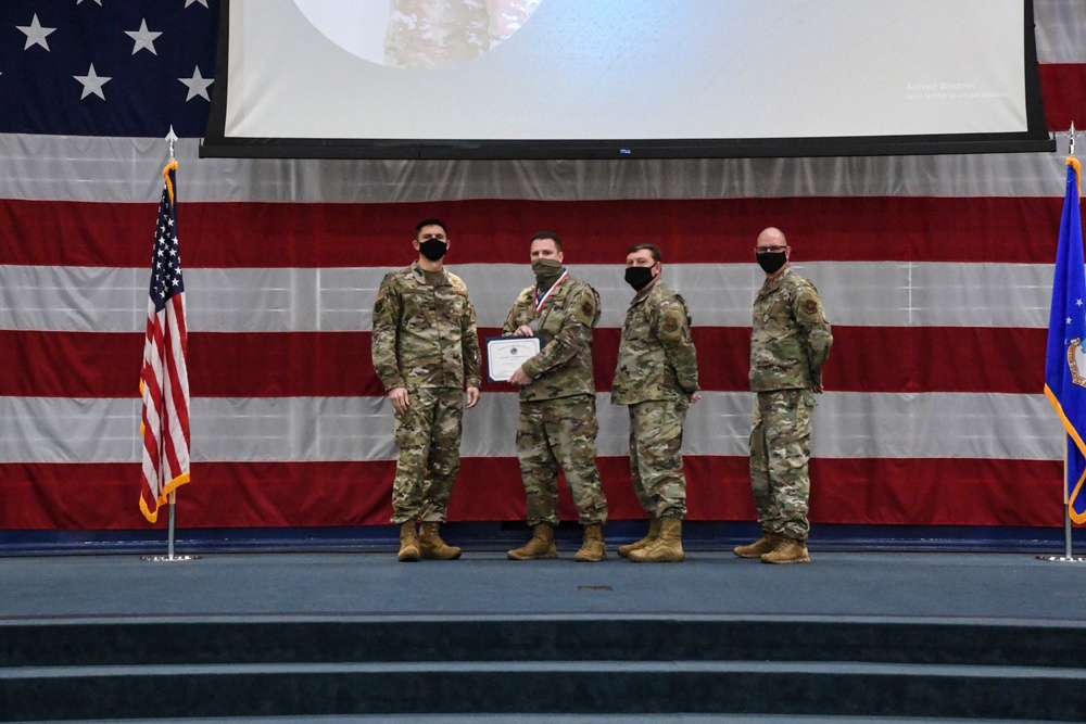 Four military personnel in camouflage uniforms stand on a stage during a formal awards ceremony. One individual wearing a medal is receiving a certificate while shaking hands with another service member, flanked by two others. The backdrop features a large American flag and a projection screen, with an Air Force flag visible on the right.