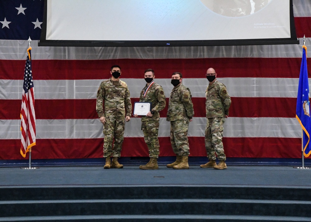 Four military personnel in OCP (Operational Camouflage Pattern) uniforms stand on a stage during a formal ceremony. One individual in the center is holding a certificate/award and wearing a medal around their neck. The backdrop features a large American flag and a US Air Force flag, with a projection screen visible above.