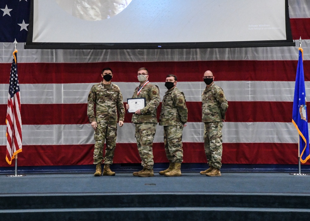 Four military personnel in OCP (Operational Camouflage Pattern) uniforms stand on a stage during a formal awards ceremony. One individual is receiving a certificate and wearing a medal/medallion around their neck. The backdrop features a large American flag drape and a projection screen, with a US flag and what appears to be an Air Force flag flanking the group.
