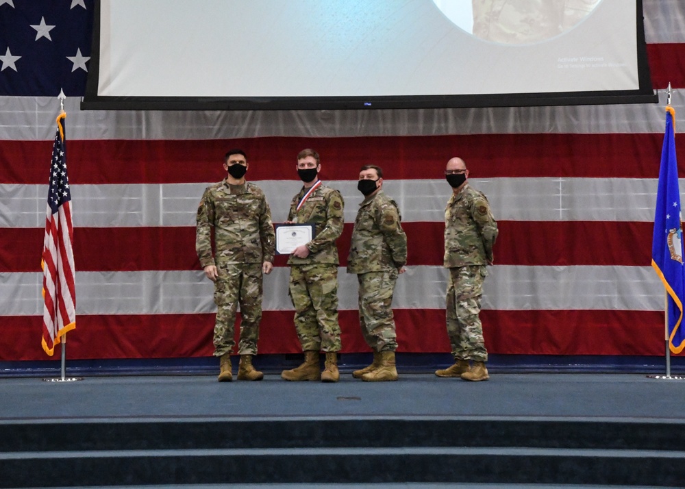 Four military personnel in camouflage uniforms stand on a stage during a formal awards ceremony. One individual is holding a certificate and wearing a medal/ribbon around their neck, indicating they are the recipient of an award. All four individuals are wearing black face masks, suggesting this event took place during the COVID-19 pandemic period.