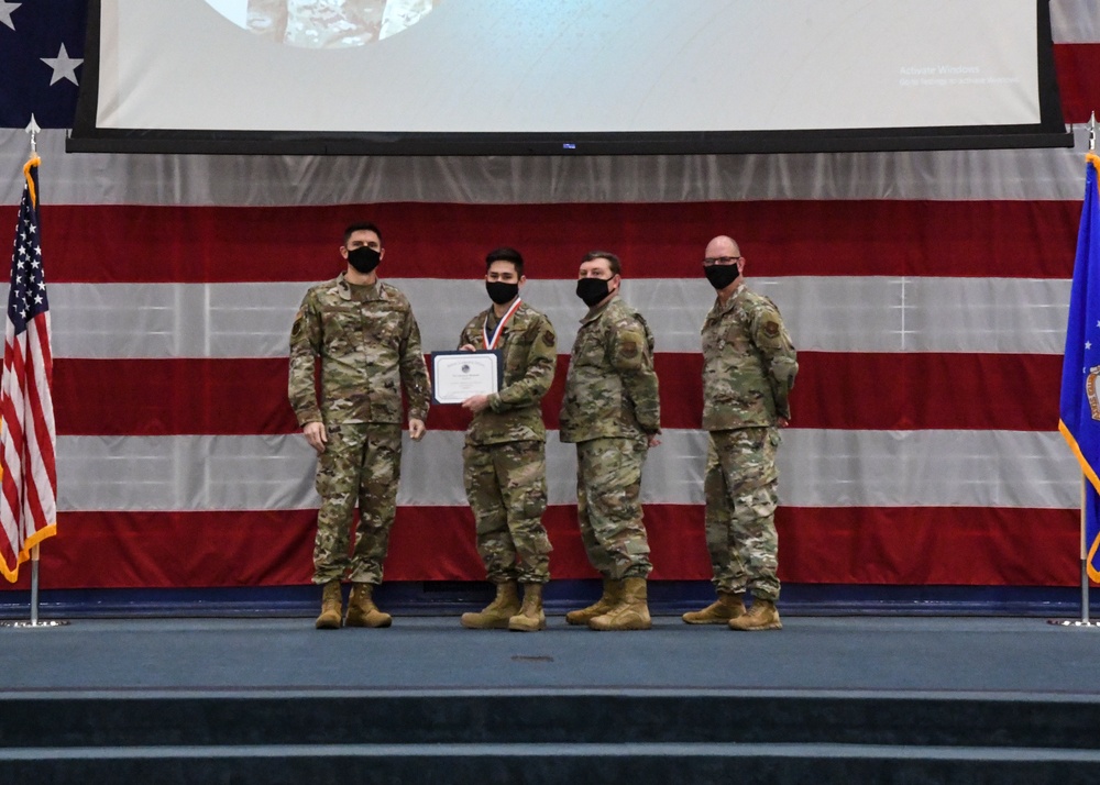 Four military personnel in OCP (Operational Camouflage Pattern) uniforms stand on a stage during what appears to be an awards ceremony. One service member is receiving a certificate and wearing a medal, while the others stand in formation. The backdrop features a large American flag and a projection screen.