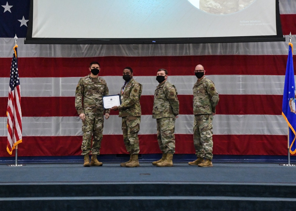 Four military personnel in OCP (Operational Camouflage Pattern) uniforms stand on a stage during a formal awards ceremony. One service member is receiving a certificate/award while wearing a medal, with two others presenting it and a fourth standing at attention. The ceremony takes place in front of a large American flag backdrop and a US Air Force flag.